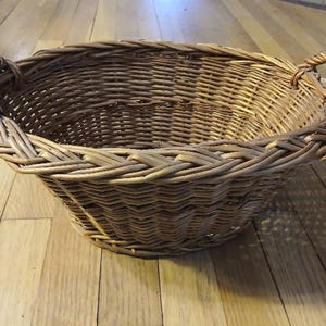 May include: A round, woven wicker basket with two sturdy handles. The basket is a warm brown color and appears to be handmade. It sits on a wooden floor.