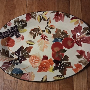 May include: An oval ceramic serving platter with a cream-colored background featuring a painted pattern of autumn leaves in shades of red, orange, brown, and yellow, along with clusters of dark purple grapes and bright red apples.