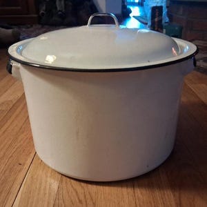 May include: A large white enamel stockpot with a matching lid. The pot features a smooth white finish, a black rim around the top edge, and two side handles. The lid has a small metal handle on top. The construction is simple and functional.