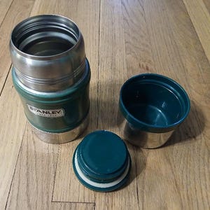 Puede incluir: Un tarro de comida Stanley de color verde oscuro y plateado con la tapa retirada y colocada al lado. El recipiente cuenta con un interior met&aacute;lico, un borde roscado y un tap&oacute;n de pl&aacute;stico verde oscuro con una tapa aislante a juego.