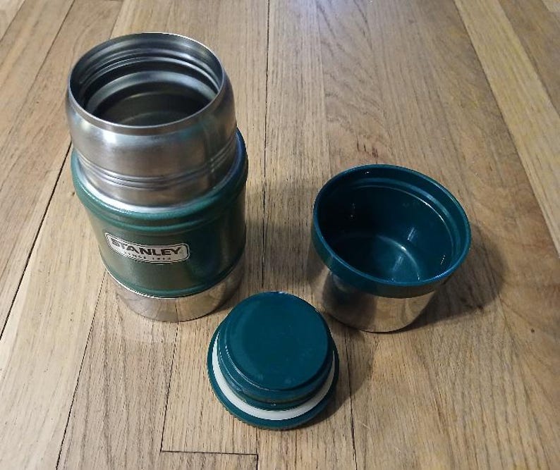 Puede incluir: Un tarro de comida Stanley de color verde oscuro y plateado con la tapa retirada y colocada al lado. El recipiente cuenta con un interior met&aacute;lico, un borde roscado y un tap&oacute;n de pl&aacute;stico verde oscuro con una tapa aislante a juego.
