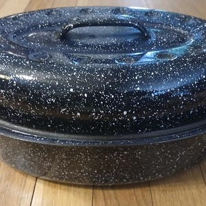 May include: An oval-shaped black roasting pan with a speckled white pattern throughout the exterior. The roaster features a matching lid with a central handle and integrated side handles on the base for lifting and carrying the cookware.
