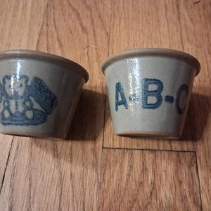 May include: Two small, light gray stoneware bowls with a smooth, glazed finish. One bowl features a dark blue stamped graphic of a teddy bear, while the other bowl displays the dark blue printed letters A-B-C on its exterior side.