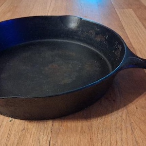 May include: A round black cast iron skillet with a long handle and a pour spout on the side. The interior surface of the pan shows a dark, seasoned finish with subtle variations in texture and color across the bottom and the curved side walls.