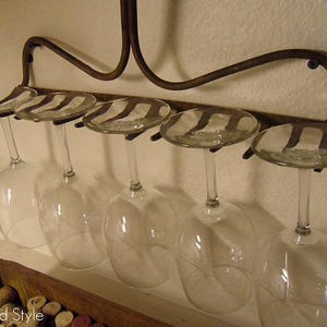 May include: A rustic, wrought-iron wine glass rack mounted on a wall, holding six clear wine glasses. The rack has a curved design and is attached to the wall with screws. The glasses are suspended upside down, ready for use.