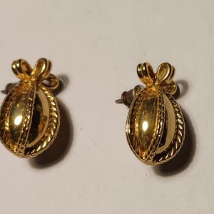 May include: A pair of gold-colored earrings. Each earring features a bow-shaped top and an oval-shaped body with a textured, rope-like border. The earrings have a post backing.