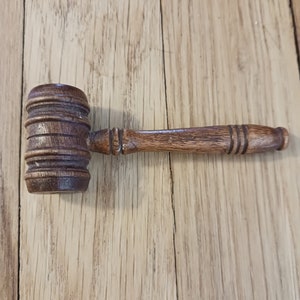 May include: A wooden gavel with a dark brown finish. The head of the gavel is cylindrical with several carved rings. The handle is slender and also made of wood. The gavel is resting on a light brown wooden surface.