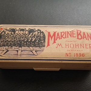 May include: A rectangular beige harmonica case with a black and white illustration of a band on the left side. The text reads Marine Band, Made by M. Hohner, Germany, and No 1896 in red and black lettering. The case has a thin blue border.