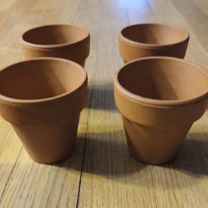 May include: Four terracotta-colored flower pots. Each pot has a slightly flared rim and a tapered body. The pots are arranged on a wooden surface, showcasing their simple, classic design. Ideal for small plants or herbs.