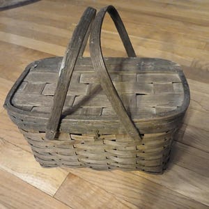 May include: A weathered, rectangular picnic basket with a hinged lid and a wooden handle. The basket is made of woven wood strips, with a natural, aged appearance. The handle arches over the top.