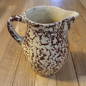 May include: A ceramic pitcher with a cream-colored base and a brown speckled pattern. The pitcher has a handle and a spout for pouring. The design is reminiscent of vintage kitchenware.