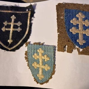 May include: Three vintage shield-shaped patches. One is black with a gold cross, another is blue with a gold cross, and the third is a smaller light blue patch with a gold cross. All have frayed edges.