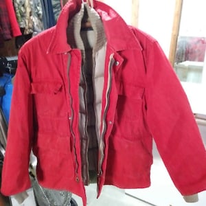 May include: A bright red jacket featuring a fold-down collar, long sleeves with ribbed cuffs, and two chest pockets. The jacket has a front zipper closure and an inner beige quilted lining that is visible through the open front panels.
