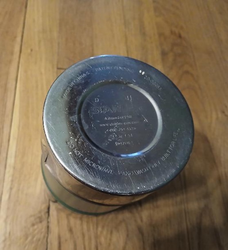 Puede incluir: La base circular de un vaso de metal plateado con texto en relieve que dice Stanley, A Brand of PMI, Made in China, Patent Pending, 20-00085, 14 oz / 0.4 L, Do Not Microwave, Hand Wash Only y Built for Life.