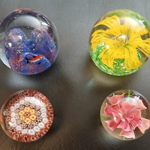May include: Four spherical glass paperweights. One features a blue and purple nebula-like interior with a small orange and black fish. Another contains a yellow flower over green foliage. A third has a complex geometric millefiori pattern. The fourth displays a pink flower.