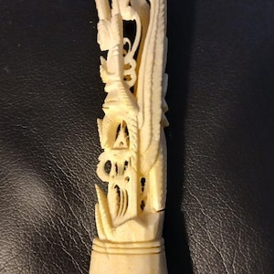 May include: Ivory-colored carved sculpture depicting a detailed figure with a spear, intricate architectural elements, and a stylized tail. The sculpture is mounted on a pedestal base, showcasing fine craftsmanship.