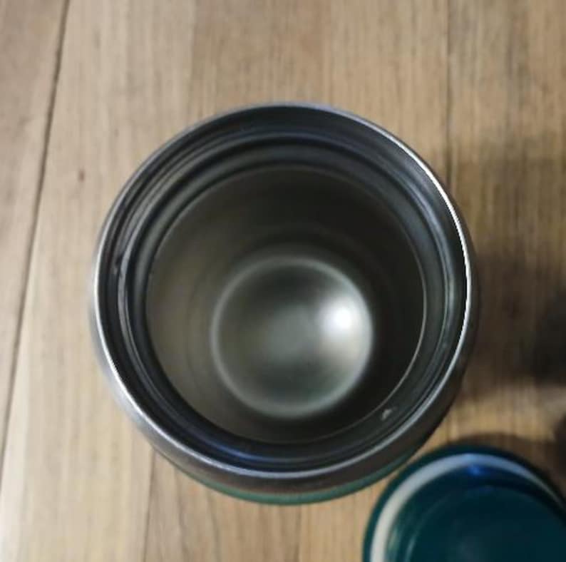 Puede incluir: Una vista desde arriba de un vaso cil&iacute;ndrico abierto de acero inoxidable con un interior de color plata met&aacute;lica. El borde del recipiente es liso y circular, mostrando la profundidad del vaso. Una tapa verde oscuro es visible.