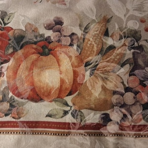 May include: A beige fabric tablecloth featuring a woven harvest pattern of orange pumpkins, red apples, green leaves, and clusters of purple grapes. The design includes a decorative border with thin red and brown stripes along the bottom edge.