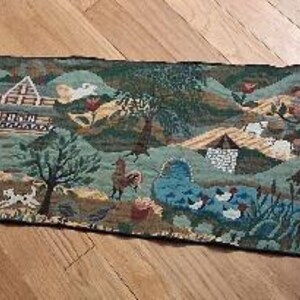 May include: A long, rectangular woven table runner featuring a detailed pastoral scene with houses, trees, chickens, and dogs in shades of green, blue, and brown. The runner has pointed ends finished with dark tassels at each of the two corners.