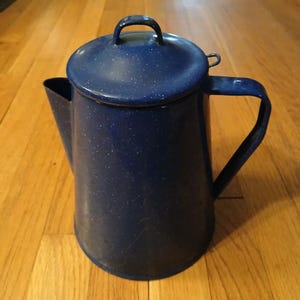 May include: A dark blue enamel coffee pot with a speckled white pattern, featuring a matching lid with a small handle, a wide spout, and a sturdy side handle. The cylindrical body tapers slightly toward the top, showcasing a classic design.