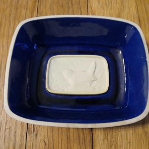 May include: A square, ceramic dish with a deep blue interior and a white rim. The center features a raised, rectangular section with a white relief design. The dish is likely for serving or display.