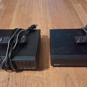 May include: Two black Sanyo DVD players, each featuring a rectangular chassis with a front-loading disc tray, control buttons, and the Sanyo logo. Each unit includes a black remote control and a power cord, with one unit featuring attached RCA cables.