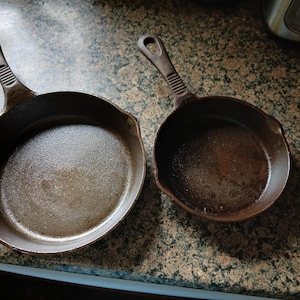 May include: Two cast iron skillets, one large and one small, sit side-by-side. Both pans are dark brown with textured cooking surfaces and attached handles. The handles have a hole at the end for hanging. The skillets are on a speckled countertop.