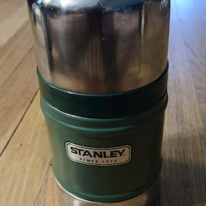 Puede incluir: Un recipiente cil&iacute;ndrico para alimentos Stanley con cuerpo texturizado de color verde oscuro y tapa y base de acero inoxidable plateado pulido. El frente presenta una etiqueta rectangular blanca con el texto negro Stanley Since 1913.