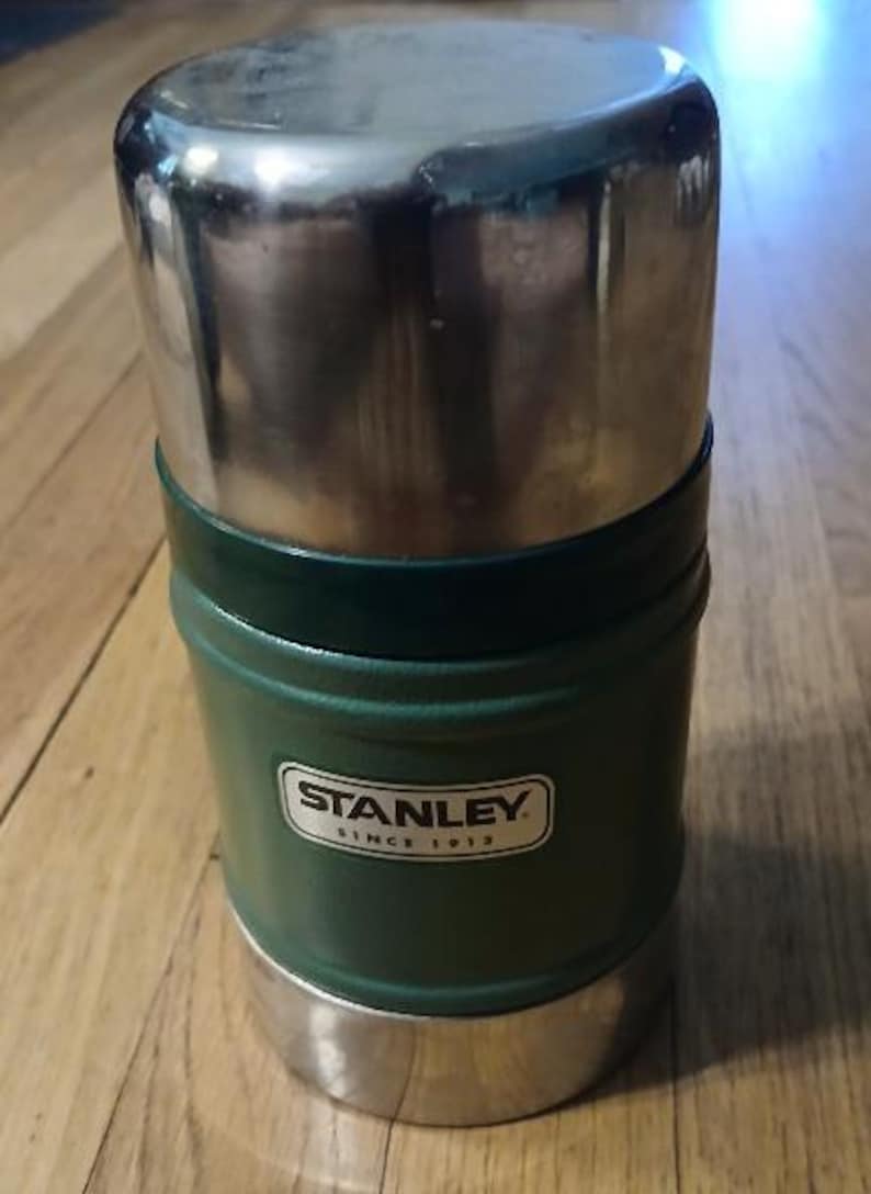 Puede incluir: Un recipiente cil&iacute;ndrico para alimentos Stanley con cuerpo texturizado de color verde oscuro y tapa y base de acero inoxidable plateado pulido. El frente presenta una etiqueta rectangular blanca con el texto negro Stanley Since 1913.