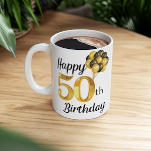 May include: White ceramic mug with a black handle. The mug has a gold number 50 with the word "Happy" above it and "th Birthday" below it. There are black and gold balloons with white polka dots on the mug.