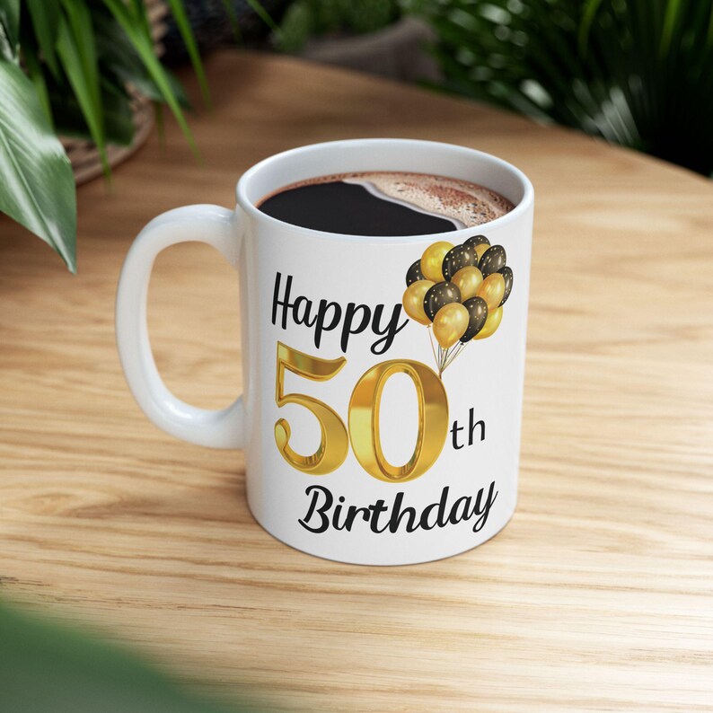 May include: White ceramic mug with a black handle. The mug has a gold number 50 with the word "Happy" above it and "th Birthday" below it. There are black and gold balloons with white polka dots on the mug.