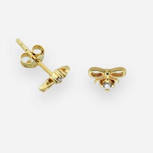 May include: A pair of gold-coloured stud earrings. Each earring features a bow design with a small, clear gemstone at the centre. The earrings are set against a plain, light background.