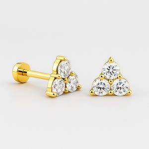 May include: Gold-colored stud earrings with a triangle design. Each earring features three clear, round gemstones. The earrings have a post and flat backing for secure wear. The jewelry is set against a white background.
