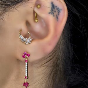 May include: A pair of gold earrings with pink and clear gemstones. The earrings are dangling and have a long, thin design. The earrings are worn in the earlobe and helix piercing.
