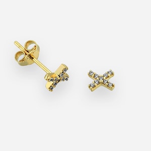 以下が含まれることがあります： 小さな透明な宝石がちりばめられた、X字型のゴールドスタッドピアス。ピアスにはポストとバタフライバックが付いています。背景は無地の明るい色です。