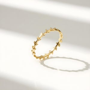Könnte beinhalten: Ein goldfarbener Ring mit einem sich wiederholenden Blattmuster. Der Ring ist vor einem weißen Hintergrund platziert, wobei Schatten Tiefe verleihen. Das Design des Rings ist ein durchgehendes Muster aus stilisierten Blättern.