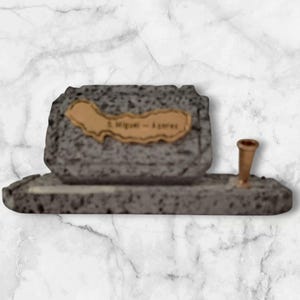 May include: A grey stone-effect sculpture with a tan plaque reading "S. Miguel - Açores". A small, bronze-coloured object is attached to the base. The sculpture sits on a marble-patterned background.