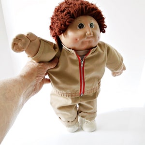 May include: A vintage Cabbage Patch Kid doll in a tan jumpsuit with a red zip. The doll has a fair complexion, rosy cheeks, and brown curly hair. It is wearing white shoes.