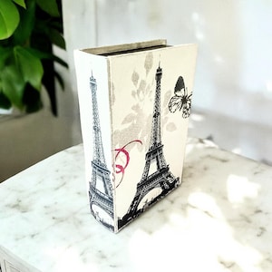 May include: A rectangular decorative box with a cream background, featuring black and white illustrations of the Eiffel Tower and a butterfly. The box has a floral pattern and is standing upright.