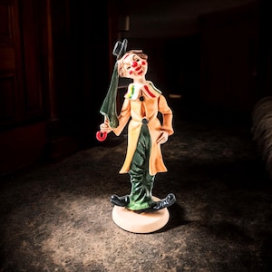 May include: A ceramic clown figurine with a tan coat, green trousers, and black shoes. The clown has a painted face with red accents and holds a green umbrella and a black top hat. The figurine stands on a round, beige base.