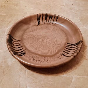 May include: A round, brown ceramic plate with a spiral design in the center. The plate has black brushstroke accents around the rim. The plate is sitting on a light brown surface.