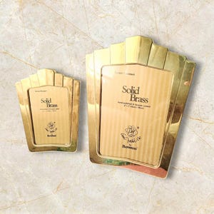 May include: Two gold-coloured solid brass picture frames. The frames have a fan-like design at the top and feature the text "Solid Brass" and "Bowon". The larger frame is approximately 7 x 10 cm.