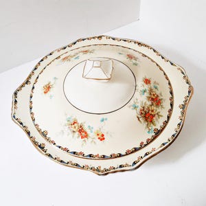 Peut inclure: Plat de service couvert de couleur crème avec garniture florale et dorée. Le plat présente un bord festonné et un couvercle avec une poignée géométrique. Des bouquets floraux rouges, bleus et jaunes ornent le plat.