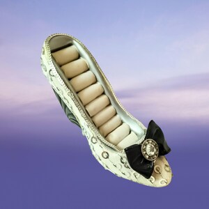 May include: A high-heeled shoe shaped jewellery organiser featuring a white exterior covered in silver sequins. The interior contains rows of cream-coloured velvet ring slots. A black bow with a central circular rhinestone embellishment sits at the toe.