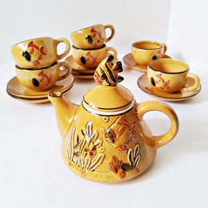 May include: A ceramic tea set with a yellow-brown glaze, featuring a teapot and several teacups with saucers. The teapot has a fish-shaped handle and lid, and the cups and teapot are decorated with fish and aquatic plant designs.