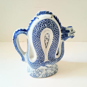 May include: White ceramic teapot with a blue dragon design. The dragon wraps around the body, with the head forming the spout. The handle and lid are also decorated with blue accents. The base features a wave pattern.
