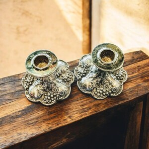 May include: Two silver candle holders with a floral and grape design. The candle holders are made of metal and have a detailed, embossed pattern. The holders are placed on a wooden surface.