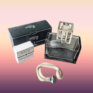 May include: A Maison Berger Paris fragrance lamp set including a dark glass vessel with a silver-toned openwork cap, a black rectangular box, a small white box labelled "BRÛLEUR POUR LAMPES BERGER," and a separate white cotton wick with a metal base.