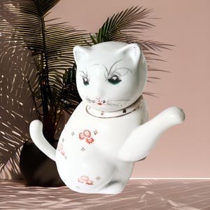 May include: A white ceramic cat figurine with a raised paw, featuring hand-painted green eyes, a gold-trimmed collar, and small red floral patterns on its body. The cat has a smooth, glossy finish and is depicted in a seated, upright posture.