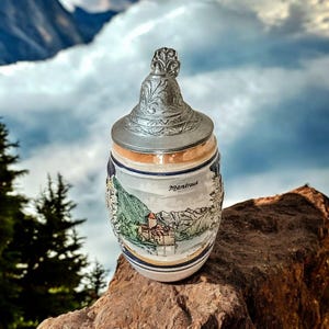 May include: A ceramic beer stein featuring a painted landscape of a castle by a lake with the text Montreux. The stein has a tapered body with blue and gold bands and is topped with a decorative, embossed grey metal lid with a pointed finial.